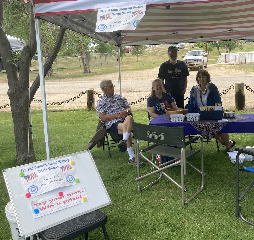 People standing near and sitting at a table under a canopy. Sign in front advertises a U.S. and Constitutional Trivia Game that awards prizes for correct answers.